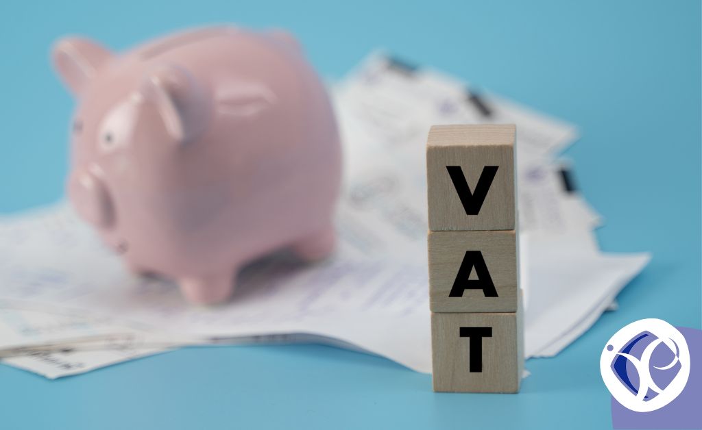 Three wooden blocks are stacked to spell the word VAT. Out of focus, behind the blocks, a cute ceramic piggy bank sits on top of some scattered documents. the image represents reclaiming VAT.